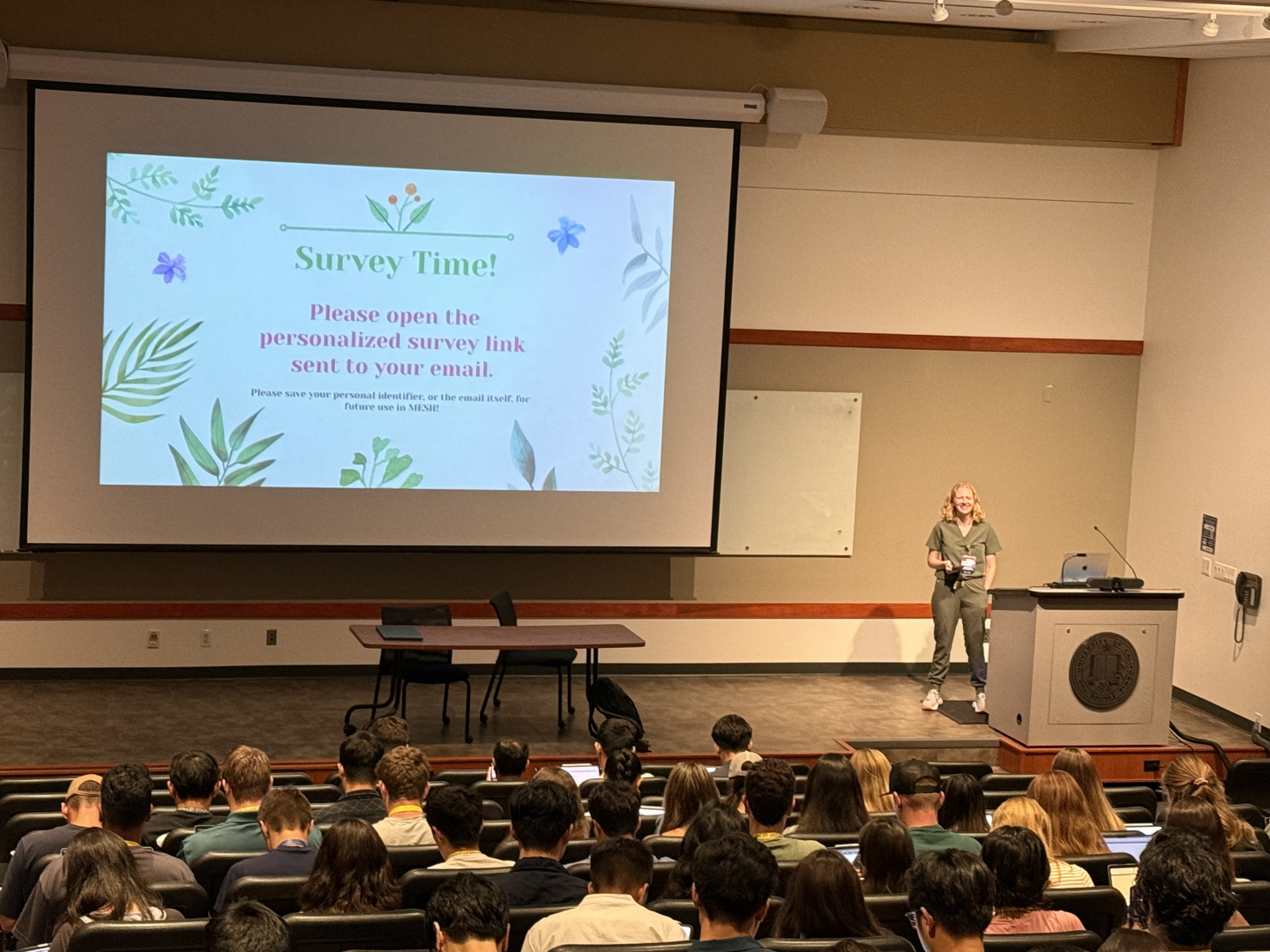 Lawrence stands at the front of a lecture hall full of students. Behind her is a large screen that reads, “Survey Time!”