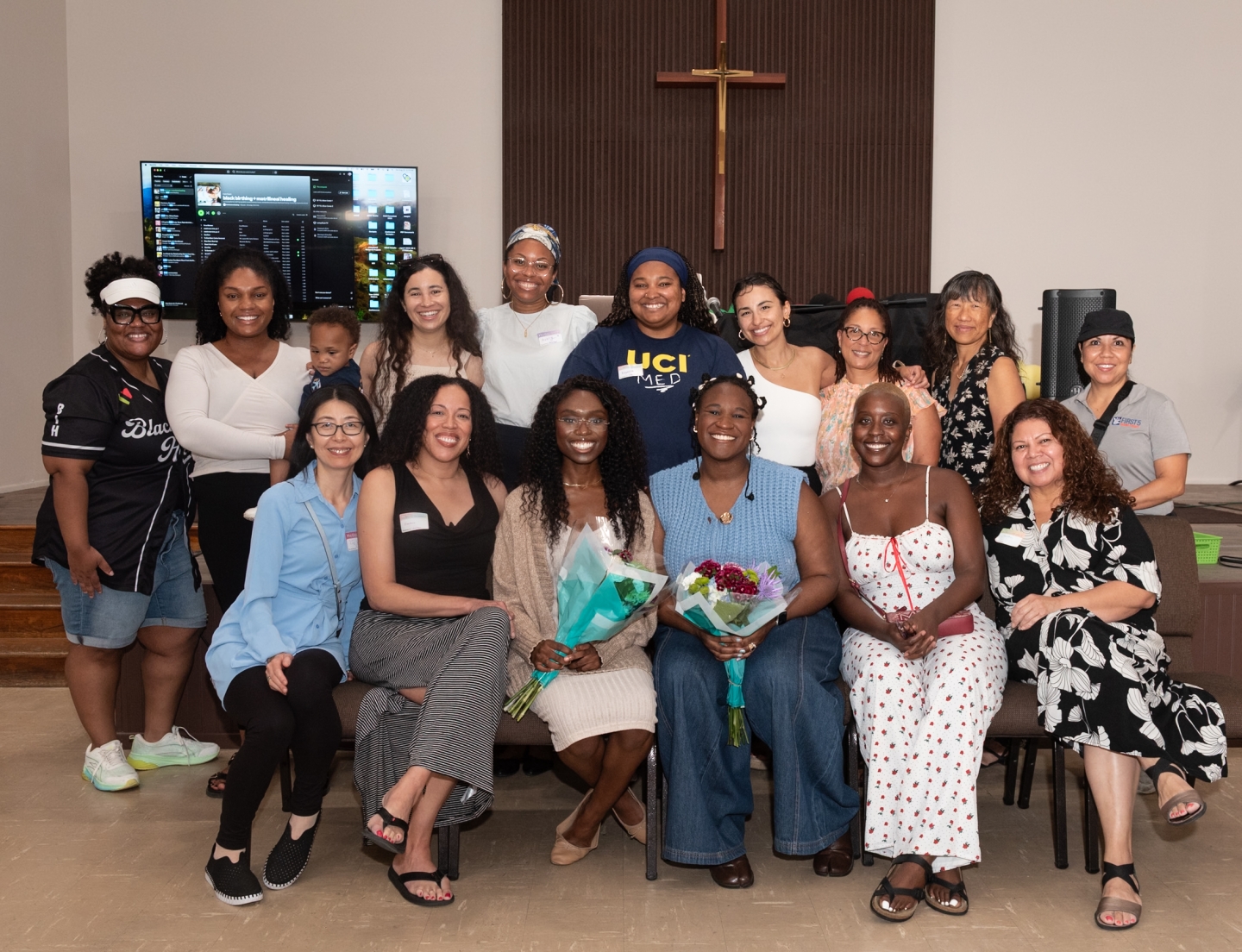 Fifteen women, one holding a baby, sit and stand together.