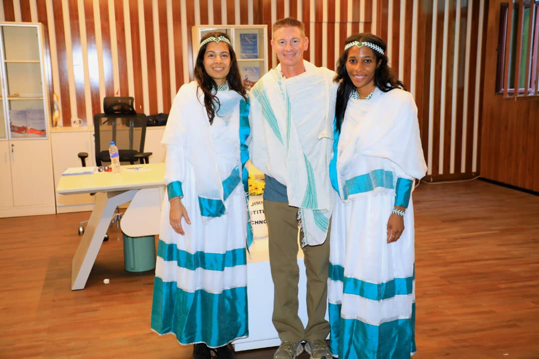 Daniel Scanderbeg stands between Priya Mitra and Chika Nwachukwu. He is draped in a white shawl with turquoise stripes. Mitra and Nwachukwu wear long white dresses with turquoise at the bottom. They’re also wearing white and green headbands and bracelets.