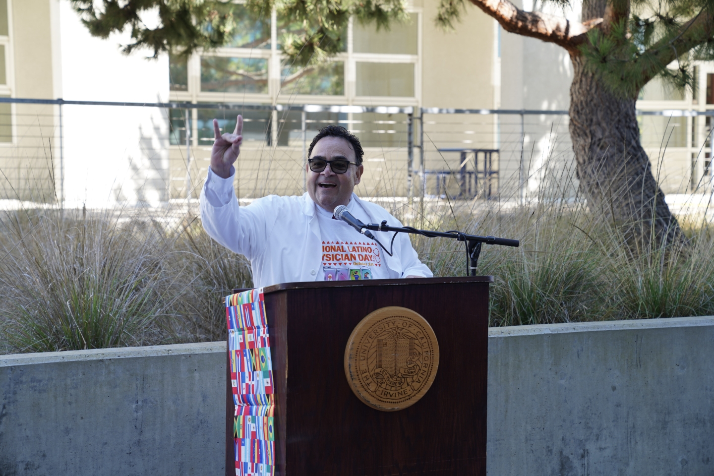 Marco stands behind a podium, giving the "zot" sign.