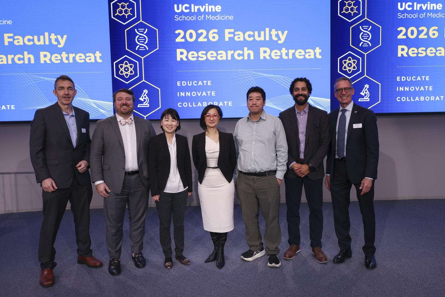 Five men and two women stand in front of two large screens that say “2026 Faculty Research Retreat.”