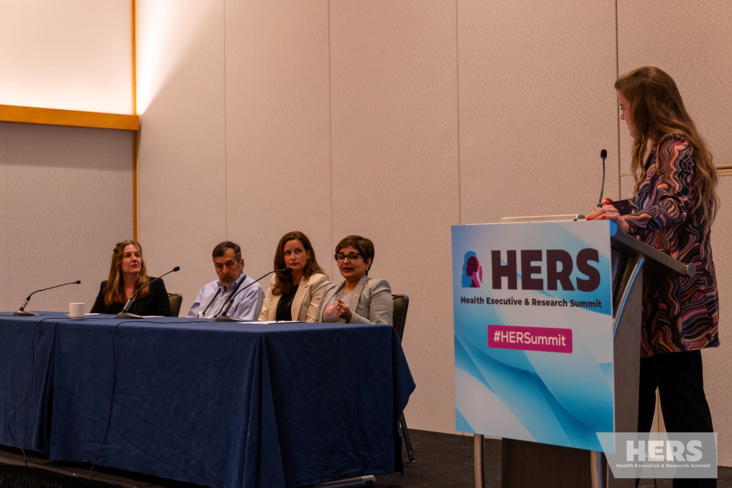 Sophie Zaaijer speaking at a podium with a sign that says, “Health Equity & Research Summit (HERS).” She is turned to the side, looking at a table with four panelists.