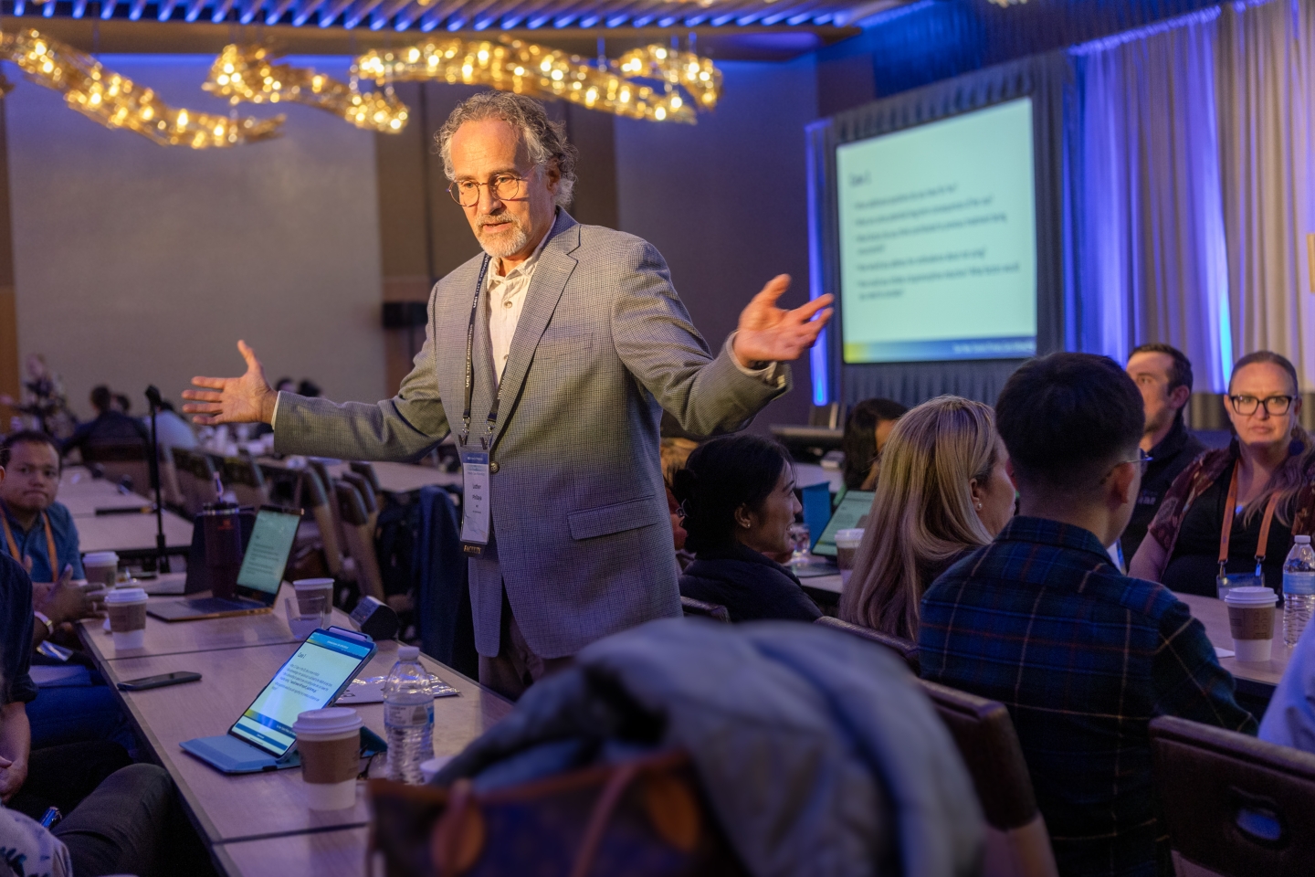 A man with gray hair and glasses gestures with open hands while speaking to an audience seated at tables. Laptops are visible on the tables.