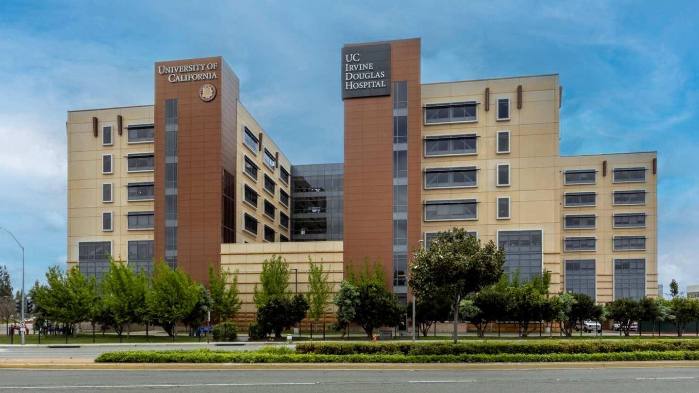 A photo of UC Irvine Medical Center — Douglas Hospital, a modern hospital building with beige walls, brown vertical panels, and rows of windows, set against a clear sky with trees and a road in front.