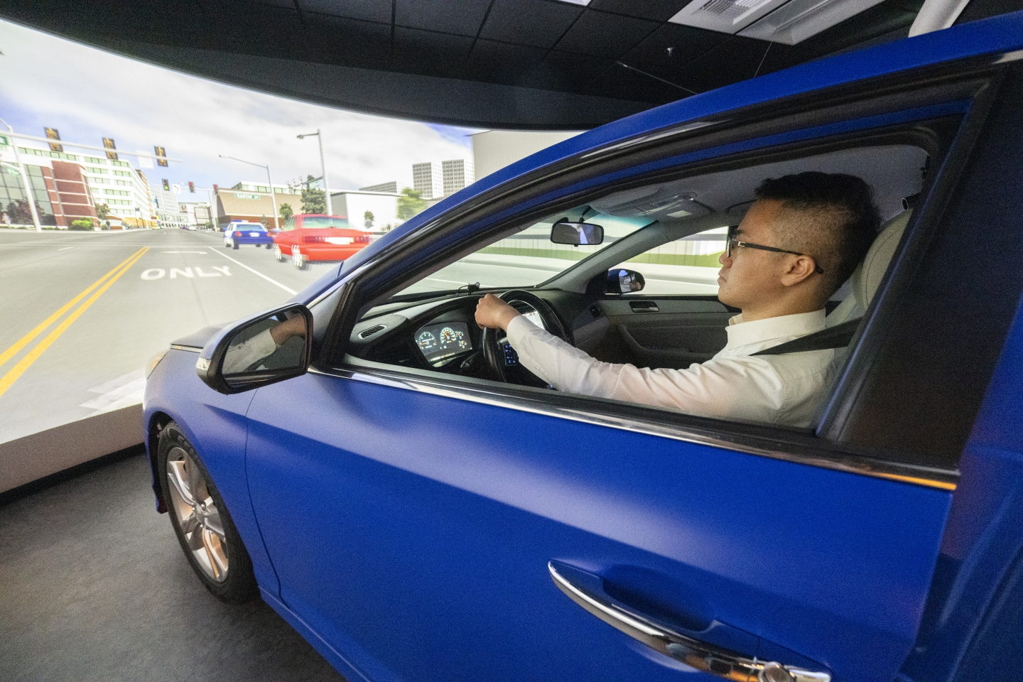 Siwei Hu sits in the driver's seat of a car in front of a screen showing a city street.