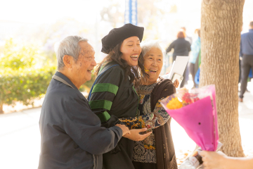 A student in her cap and gown stands smiling between an elderly couple, hugging the woman. The man is holding the student’s arm.