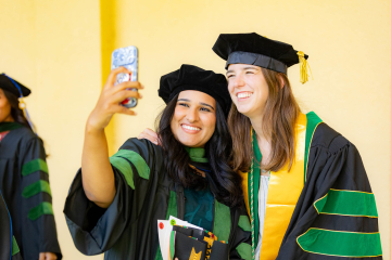 Two women in their caps and gowns smile, as one holds up a cell phone 