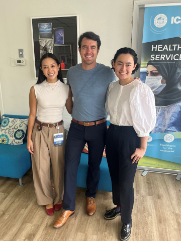 Ego, Watson and Adams stand in the clinic waiting room. Behind them is the frame collage of student artwork and a health services banner with a woman wearing a face mask.