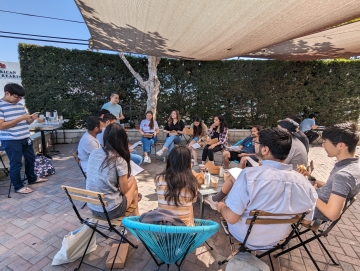 A group of people seated in a circle on a patio, engaged in a discussion under a shade cloth.