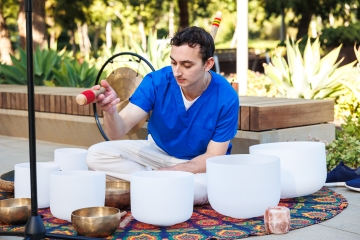 A medical student sits with a various bowls surrounding him, holding a an item for making music with them.