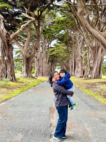 Mitul stands, holding her toddler son in her arms, on a street lined with huge trees.