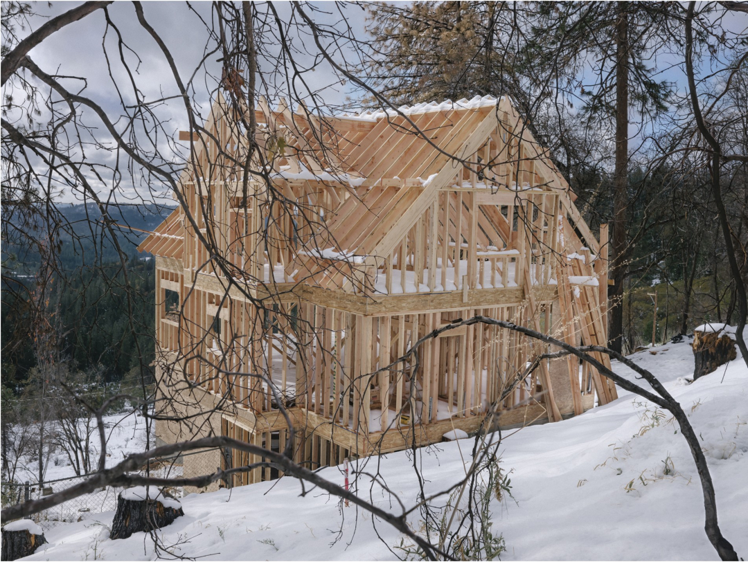 The wood frame of a house sits on a hill in the snow, with tree branches all around and mountains in the backdrop.