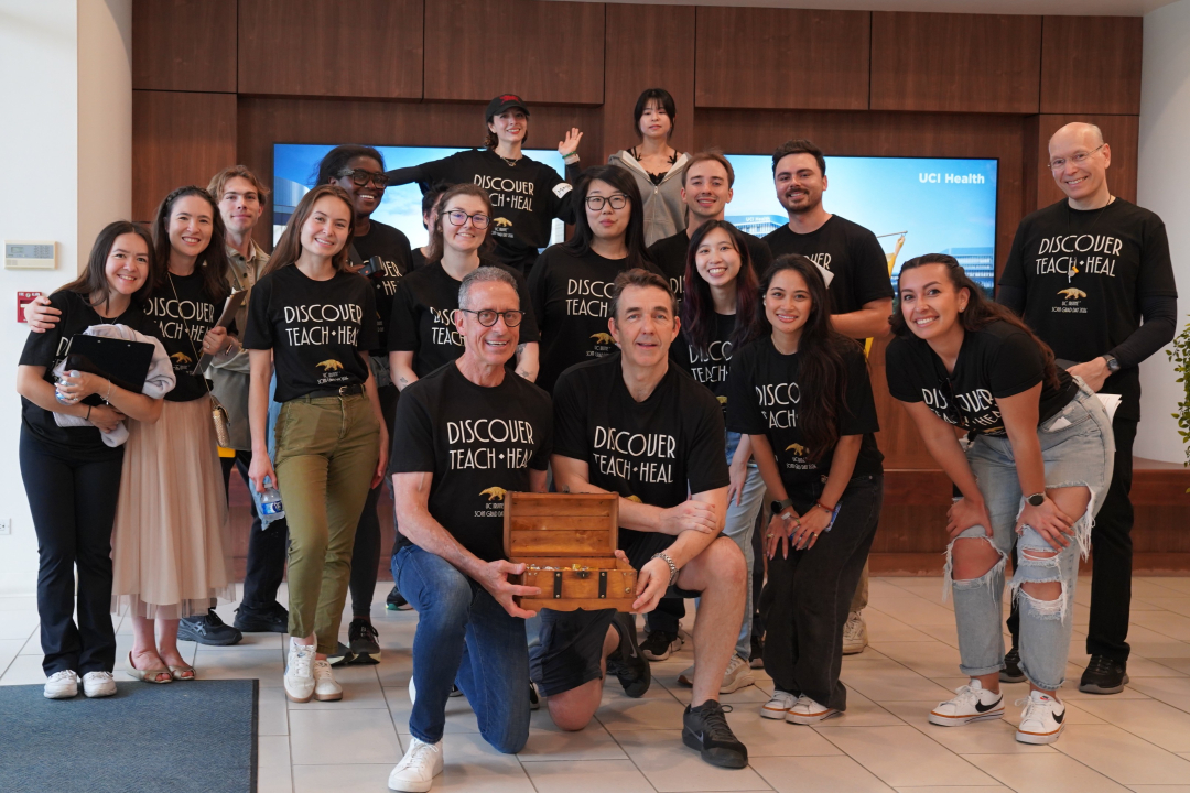 A large group of people stand behind Dean Stamos and Vice Dean Abbot, who kneel in front holding a small treasure box.